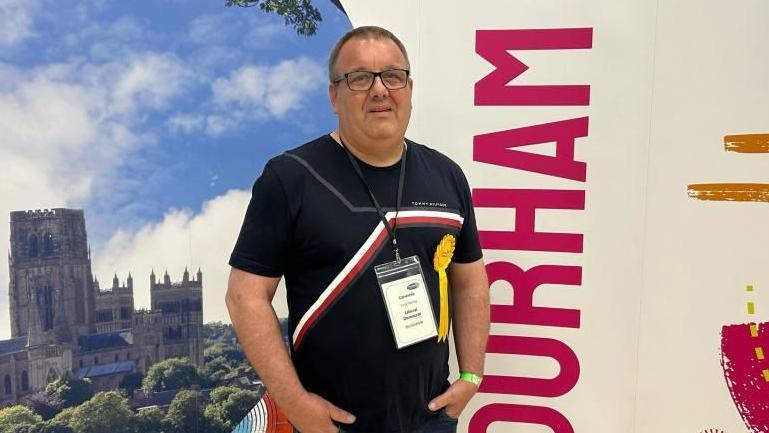 Terry Rooney smiling into the camera with his hands in the front pockets of his trousers and is standing in front of a bored which has a photo of Durham Cathedral on it. He is wearing a black top with a red and white stripe across it. He has short, dark hair and is wearing glasses.