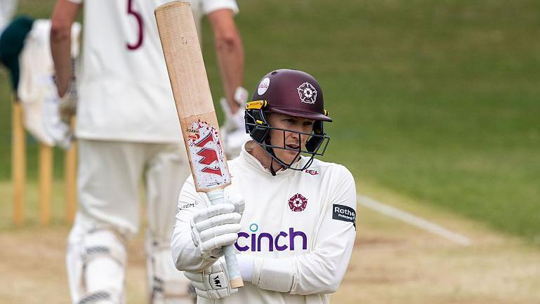 Northamptonshire batter George Bartlett, wearing white gloves, a white shirt and a maroon helmet, raises his bat