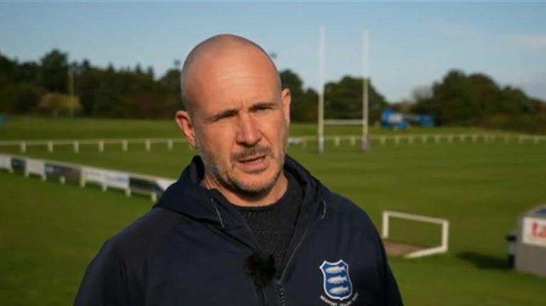 A man with close-shaven head and stubble wearing a blue club fleece and standing in front of a rugby pitch. 