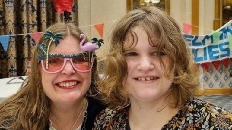 A woman and her teenage daughter smiling at the camera. The woman is wearing pink flamingo sunglasses, a dark coloured cardigan and white top. The teenager has shoulder-length curly hair and is wearing a black and orange pattered shirt. There is colourful bunting and banners in the background.