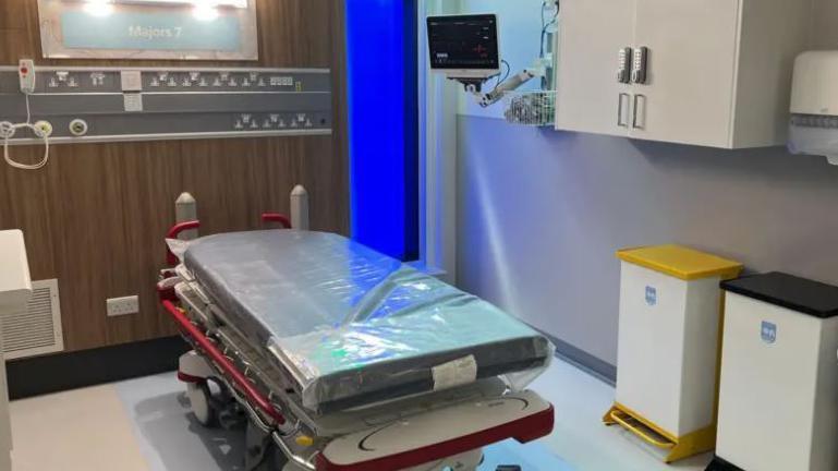 A bed in the new cubicle for emergency patients at Great Western Hospital. There is a plastic over on the bed and it's positioned near two bins.