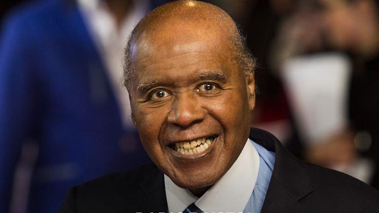 A portrait image of Paul Stephenson smiling at the camera wearing a coat, shirt and tie