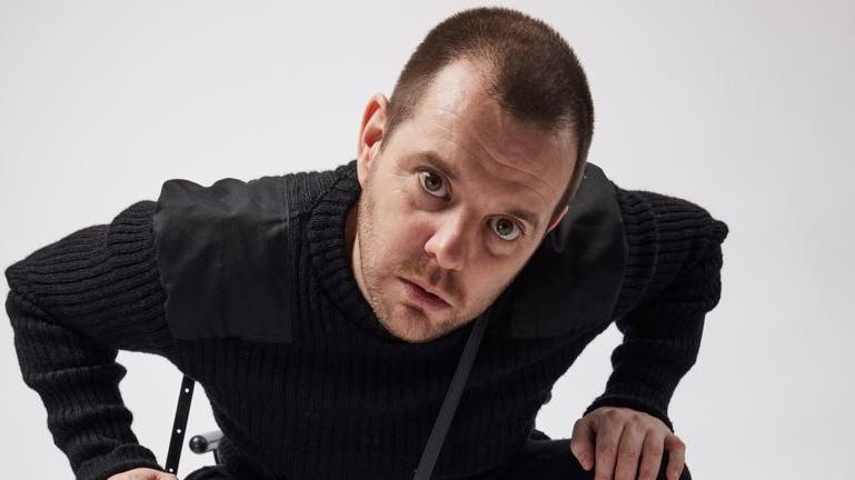 An image of Mike Skinner, front man of The Streets. He is leaning towards the camera and is wearing a black jumper.