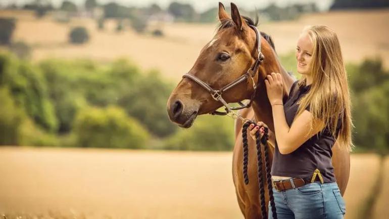 Picture of Gracie holding onto the reigns of a horse in a rural field. She had blonde hair and is wearing jeans a polo shirt. The horse is chestnut brown. 