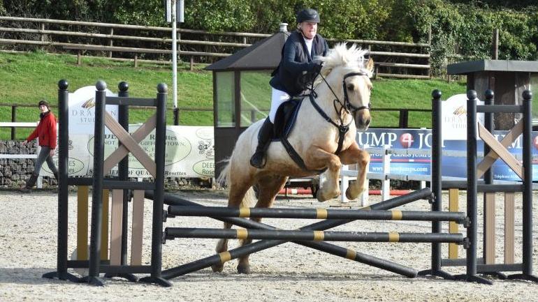 Karen is in mid-jump over a fence, made up of black cross poles, during an eventing session, riding her horse and jumping over a fence, above the showground's sand.