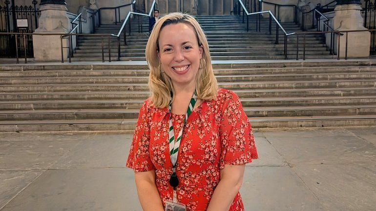 Sarah Hall MP in the Palace of Westminster. She is wearing her mid-length blonde hair down in waves, and is wearing a red flowery dress with gold hoop earrings and a lanyard around her neck. She is standing in front of stone steps.