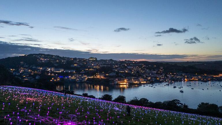 A wide angle of the Field of Light with Salcombe in the background. It is dusk and the lights are all switching on. They illuminate the entire field. There is a silhouette of a person stood in the centre of the field.