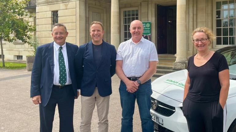 Councillor Paul Sample, with business leaders Lee Sheppard of apetito, Rob Stringer of Plastic Engineering Solutions, and Good Energy founder Juliet Davenport standing outside a house with pillars. There is a white car behind them which has 'Wiltshire Council' written on it in green writing. 
