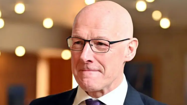 Shaven-headed John Swinney in black glasses wearing a dark suit, white shirt and purple tie