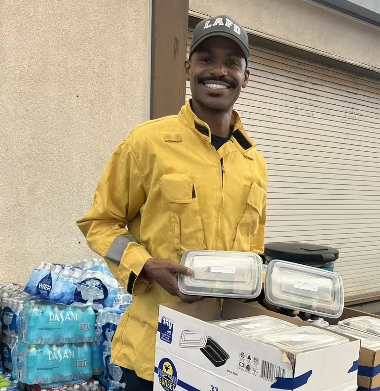 A man in a yellow jacket holds several trays of meals, as he wears an LA fire department hat, standing in front of a crate of bottled water