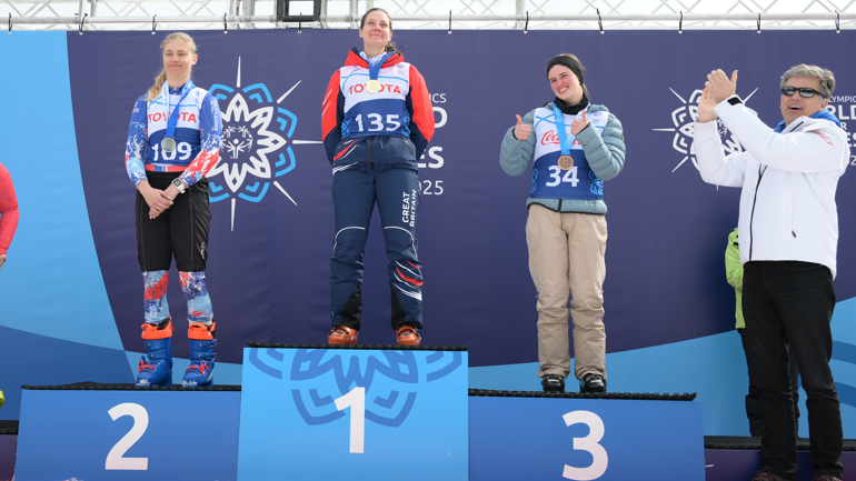 Three women in skiing gear on a blue podium with 2, 1, and 3 in white numbers below where they are standing. Each woman has a medal around her neck and is smiling. A grey-haired woman in a white jacket, sunglasses and black trousers to the right of the podium is clapping.