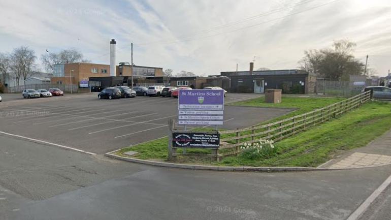 A school seen from its vehicle entrance. A purple sign for St Martins School is at the entrance, which has a wooden fence on its perimeter. Cars are parked in a car park in front of the one and two-storey buildings. There are clouds in a blue sky.