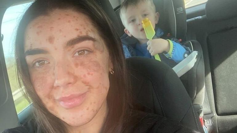 Laura Kelly is seen in the car with her son. He is in the backseat in a booster chair and she is in the passenger seat wearing black and taking a selfie.