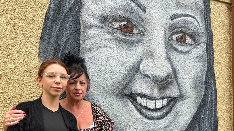 Deanna Ellis, who has light brown hair and glasses, and Sandra Bartlett, who is wearing a leopard print top and has black, standing stood in front of a wall mural of Jade Ellis