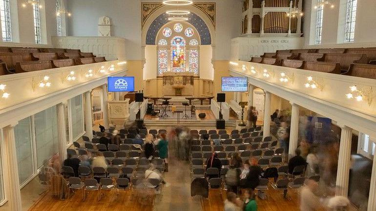 An image of St John's church in Chatham taken from upstairs. A number of people can be seen both standing and sitting, with two TV screens on either side. An altar and stain glass window can be seen at the front of the building. 