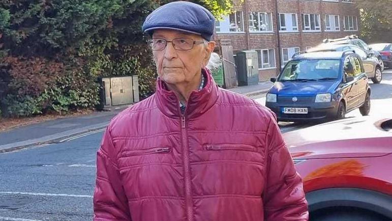 Robert Boyce in a red puffer coat looking down the road. He wears glasses and a blue flat cap.