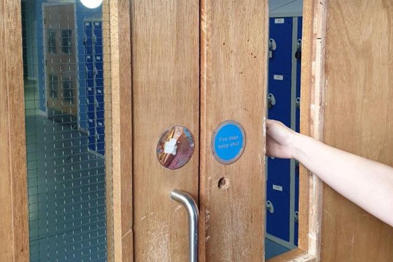 A set of double fire doors in a school, one of which has a pane of glass missing - to illustrate this a person puts their hand through the door where the glass should be