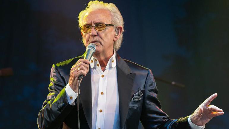 Tony Christie, who has white hair and is wearing sunglasses, wore a black suit with a white shirt 