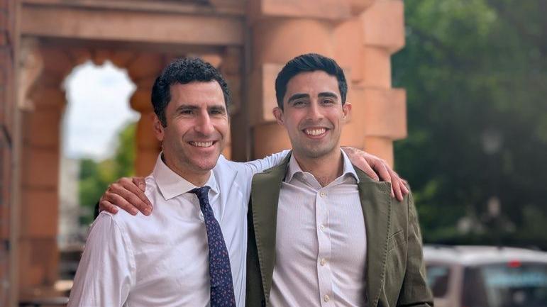 Photo of Prof Hani Marcus and Mr Danyal Khan standing outside. Both have dark brown hair. One is wearing a white shirt and blue and white tie. The other is wearing a light coloured shirt and kaki blazer.
