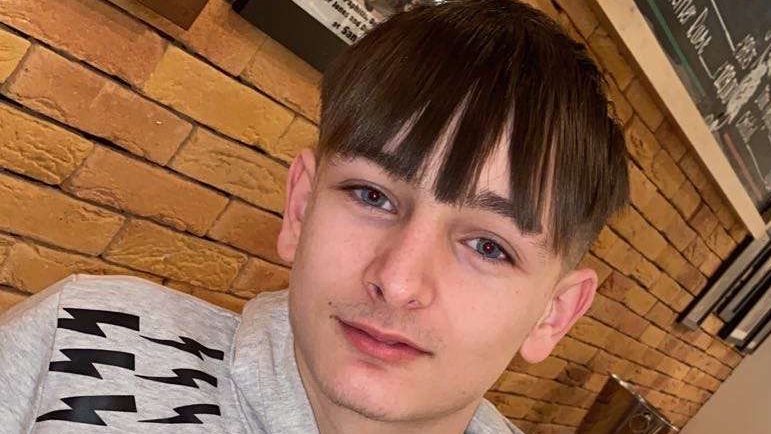 Jamie Meah looks at the camera wearing a grey hoodie. He has dark medium-length hair, with the brick wall of a bar behind him. 