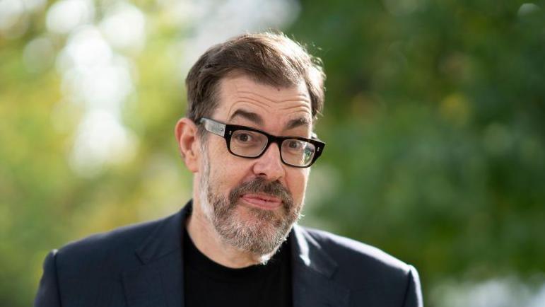 A picture of Richard Osman. He is wearing a navy suit with a black t-shirt. The background is out of focus trees.