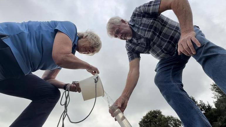 Two volunteers pour river water from a small white bucket into a clear sampling tube