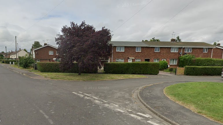 View of road junction in residential area with wide grass verges and modern red brick terraced houses behind neatly-trimmed hedges