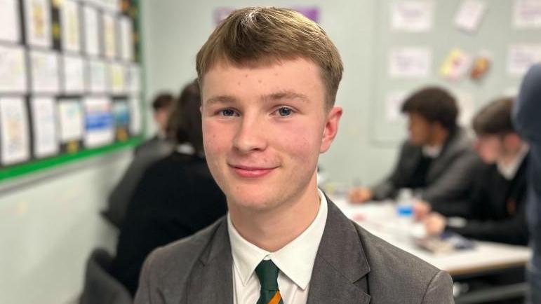 Michael has sort brown hair, he is wearing a school uniform which has a green and yellow striped tie. He is also wearing a grey blazer.