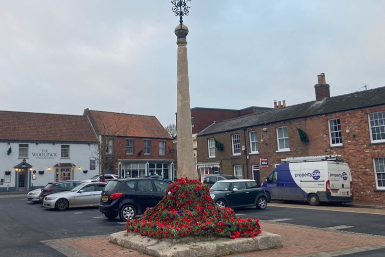 A wider view of the monument, which now stands in the middle of a carpark in the centre of the town.  Old buildings provide a backdrop to the scene.