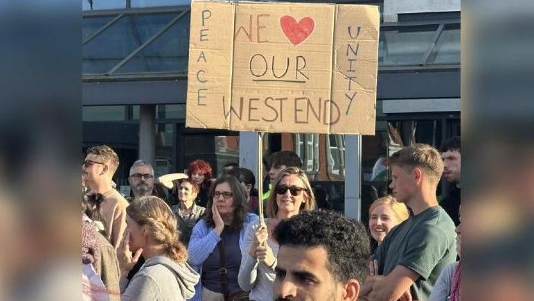 A woman holds a sign saying 'Peace, Unity, We love our West End'