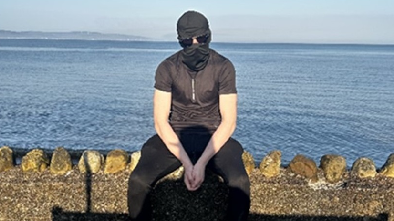 Alex Hutton, who has been jailed for more than five years, sits on a wall in front of the sea while wearing a balaclava and sunglasses