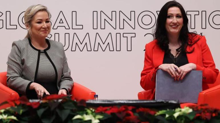 O'Neill (left) is sitting on a red chair, wearing a grey dress and matching blazer. Her blonde hair is tied up, Little-Pengelly is sitting on a red chair and is wearing a black dress and red blazer. She has long dark brown hair.