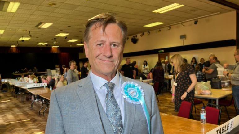 A man in a grey suit standing in an election hall. He has a light blue and white badge on he chest,