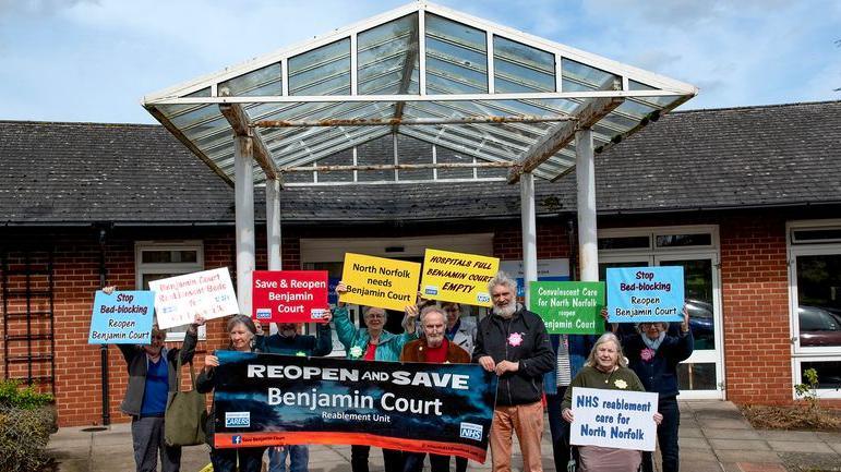 A brick building with a glass entrance with a group of protestors gathered outside holding banners reading "Save Benjamin Court"