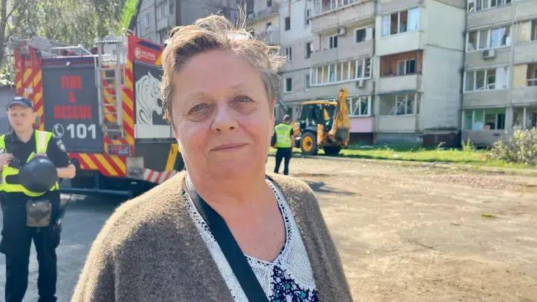 Valentina, a lady with short hair in a brown cardigan and floral shirt stood in front of the apartment building with firefighters and a truck in the background