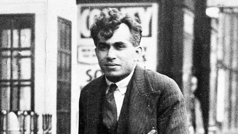 Black-and-white picture of Dr Buck Ruxton, wearing a three-piece suit and tie. He is walking next to railings and there is a row of shops in the background.