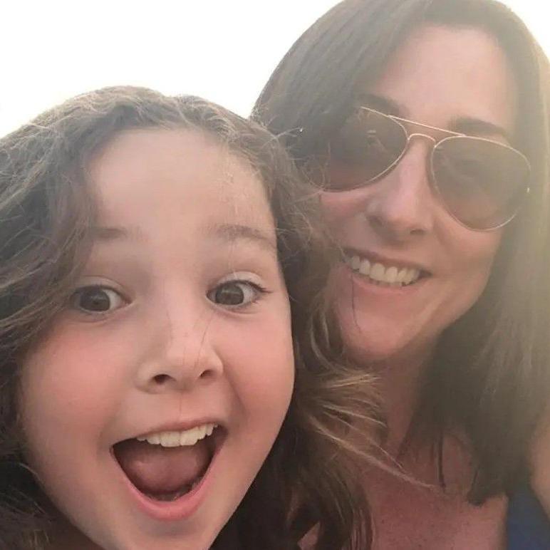 A young girl, Milly Main, with long brown hair smiles towards the camera. Her mother, Kimberly Darroch is smiling towards the camera next to her. 