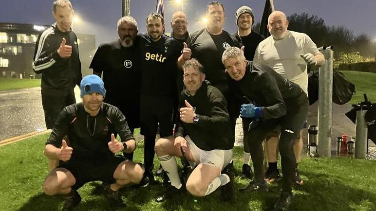 A group of men standing under streetlights on a green space. They are smiling for the camera and some of them are giving thumbs up. Two of them are kneeling in front of the group. It is dark and the road is wet. There is a building with the lights on behind them.