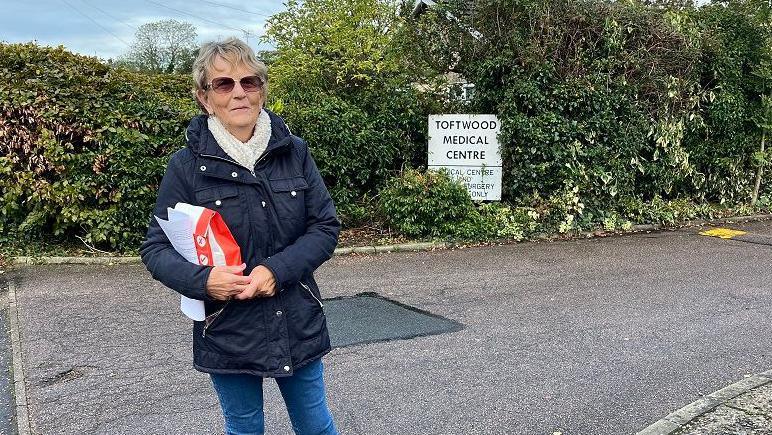 Valerie Savory standing in front of a sign saying Toftwood Medical Centre. She is wearing sunglasses, carrying a bag of prescription medication and wearing a blue coat and jeans.