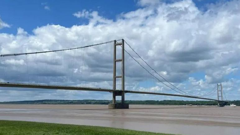 An elegant suspension bridge, with two tall concrete towers, spans the wide Humber estuary.  The water is brown and the sky blue with white clouds.