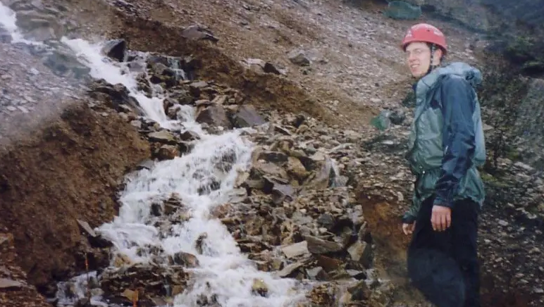 Professor Richard Johnson, wearing a red helmet, blue coat and trousers, is standing next to large rocks with water cascading down the middle of a crevice in the rock.