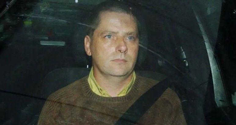 A man in a shirt and crewneck jumper gazing through the windscreen of a car. He is in the passenger seat and is wearing a seatbelt