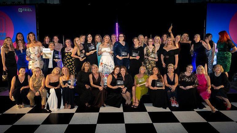 A large group of people posing together on a black-and-white checkered floor at an indoor event. Many individuals are dressed in formal or semi-formal attire, including dresses and evening wear. Several people are holding rectangular awards or plaques.