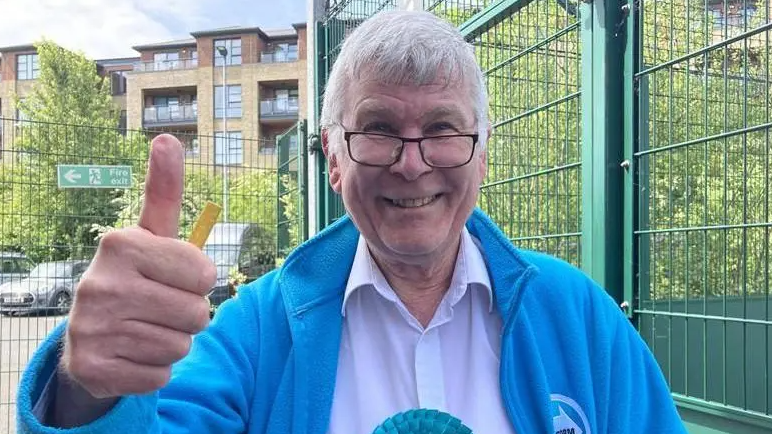 Mr Hopkins has grey hair and is wearing a blue jacket and white shirt with a blue rosette on. He is smiling and giving a thumbs up to the camera. He is stood next to a green metal fence and there is a block of flats behind him.