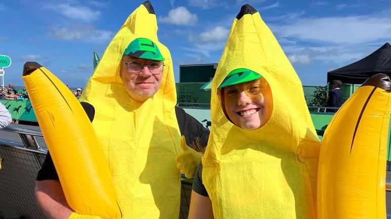 Two men dressed in banana costumes smile at the camera behind a barrier.
