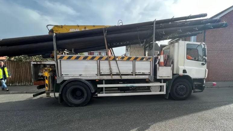 A truck is stationary and is holding several long black poles on the back of it. It is parked next to a house and you can see someone in high vis on the left.