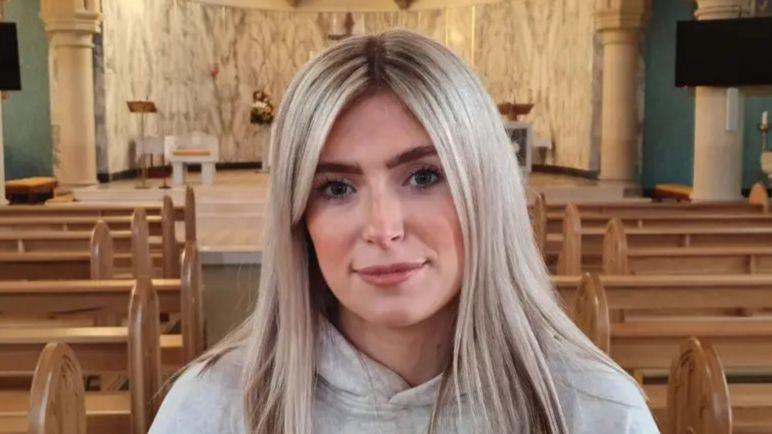 Siobhan has long, straight blonde hair. She is wearing a grey hooded sweatshirt and is looking directly into the camera. She is inside a church with rows of wooden pews in the background.