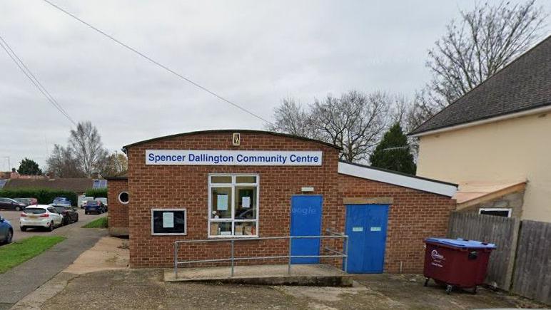 Single-storey brick building with "Spencer Dallington Community Centre" on a sign above the main window.  It has blue doors and notices in the window, with a ramp up to the main door. There is a skip to the left and cars parked in the road to the right.