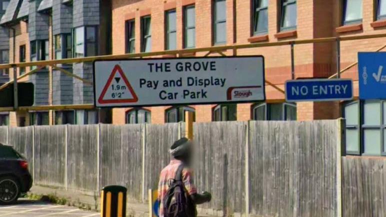 Sign for the entrance to the Grove Car Park in Slough showing height restrictions. Someone is walking past and their face is blurred out.
