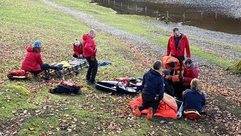 Several mountain rescue volunteers - dressed in red clothing - attend to a casualty on the path by Otterbield Bay, Derwentwater. There are lots of leaves scattered on the small slope and a stretcher has been prepared for the casualty. 